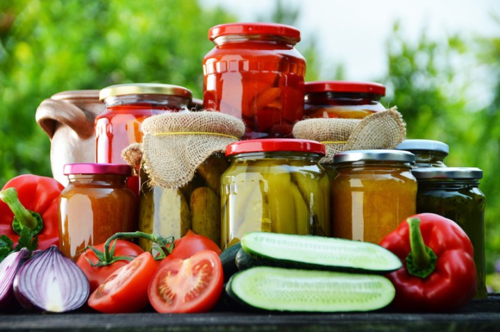 Assorted jars of homemade preserves and pickles on a rustic table, surrounded by fresh tomatoes, peppers and cucumbers.
