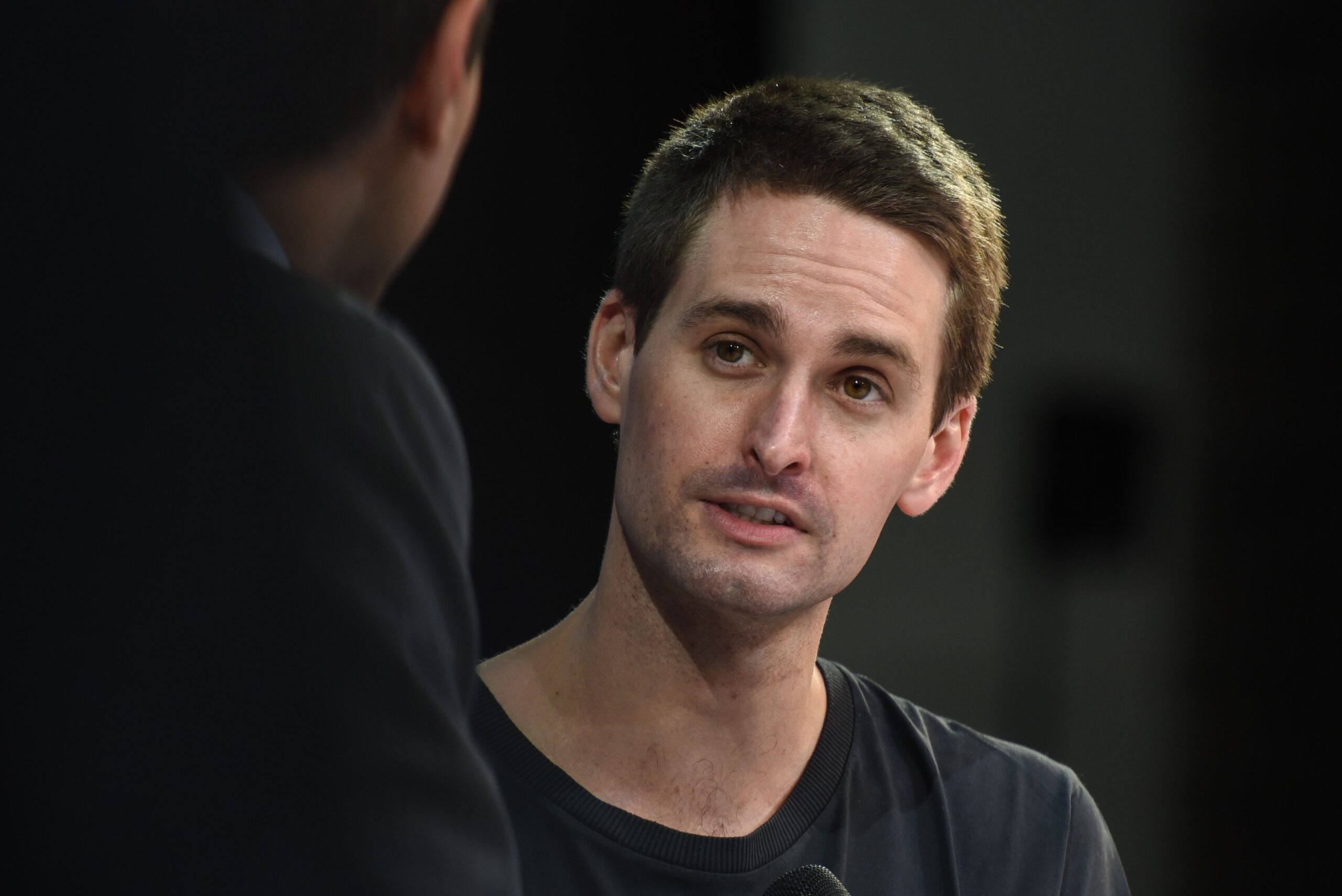 Close-up of a man with short brown hair in a black shirt, speaking during an interview, with another person partially visible in the foreground.