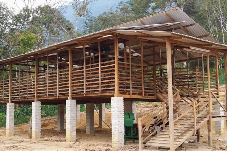 Elevated wooden building on concrete pillars with an open-slatted design and a staircase on the right under a metal roof in a rural area.