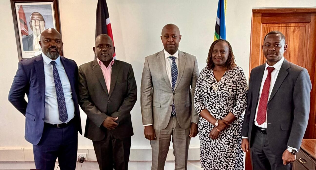 Five formally dressed adults standing in a row for a group photo in an office, with flags and a framed portrait in the background.