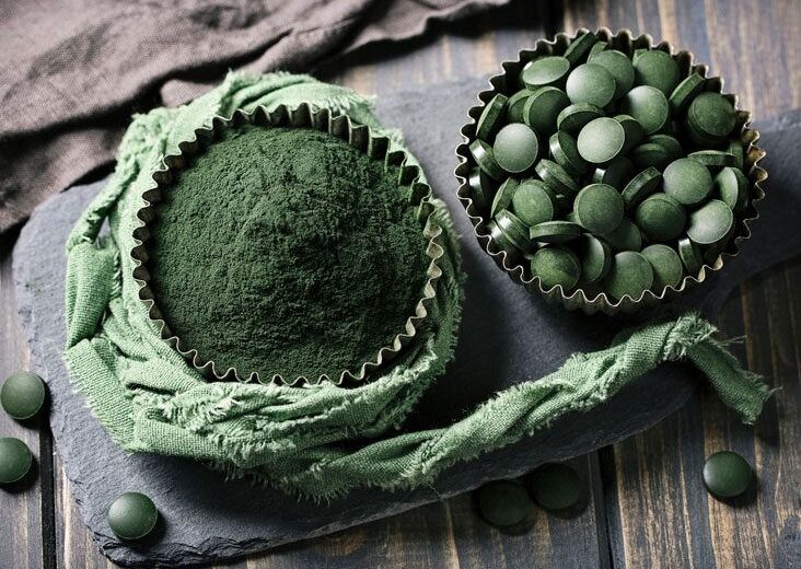 Cup of green matcha powder next to a tin of spirulina tablets on a dark slate board with a green cloth.