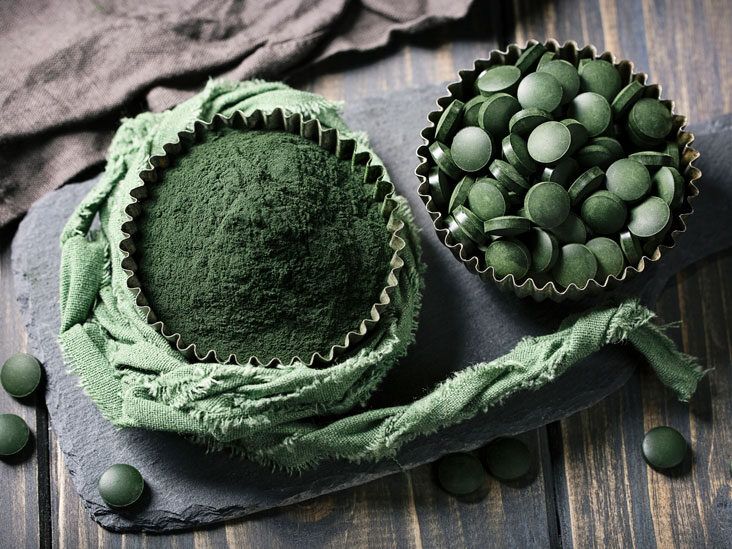 Cup of green matcha powder next to a tin of spirulina tablets on a dark slate board with a green cloth.
