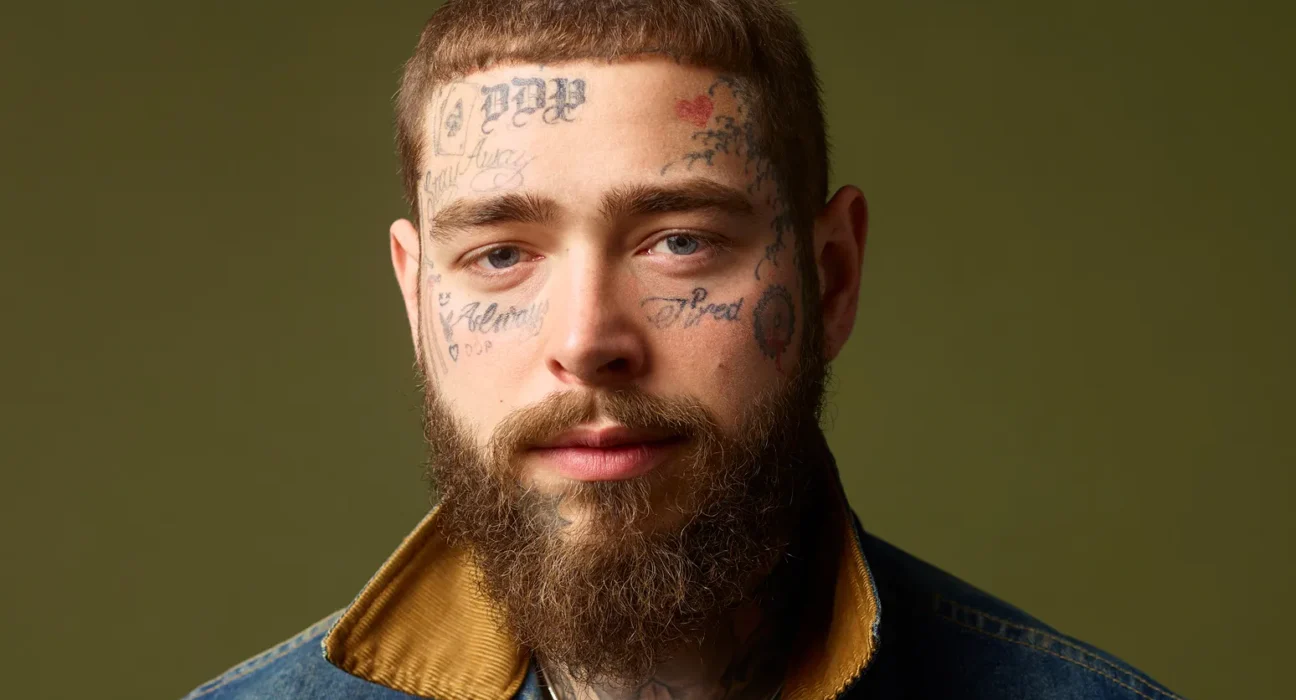 Close-up portrait of a bearded man with facial tattoos wearing a denim jacket, looking at the camera