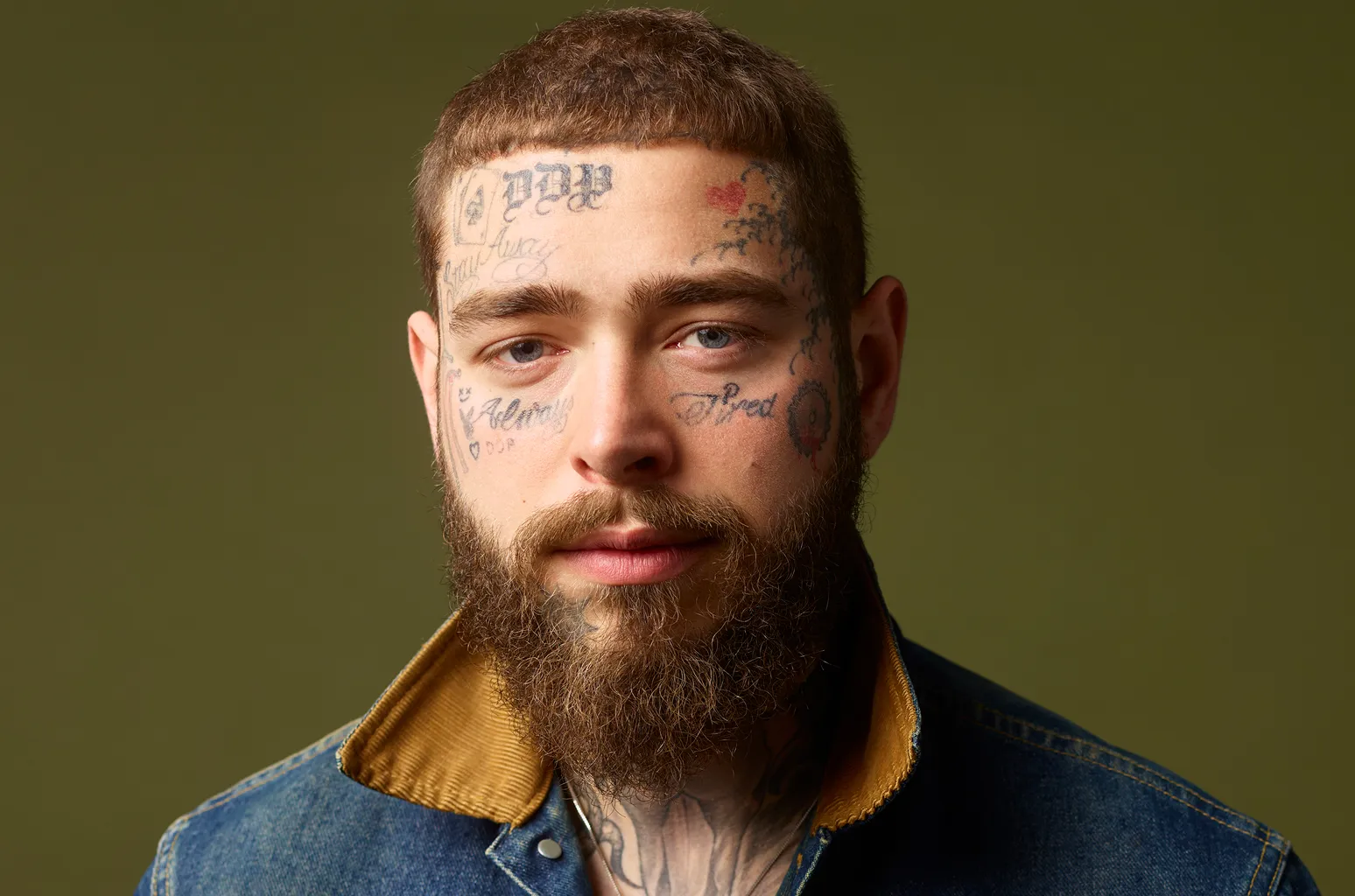 Close-up portrait of a bearded man with facial tattoos wearing a denim jacket, looking at the camera