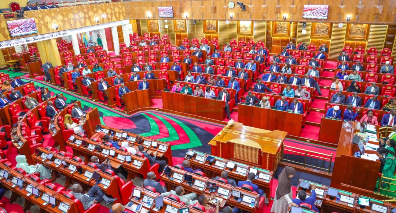 Parliament chamber filled with lawmakers seated in red chairs around wooden desks, with a central podium and colorful carpet.