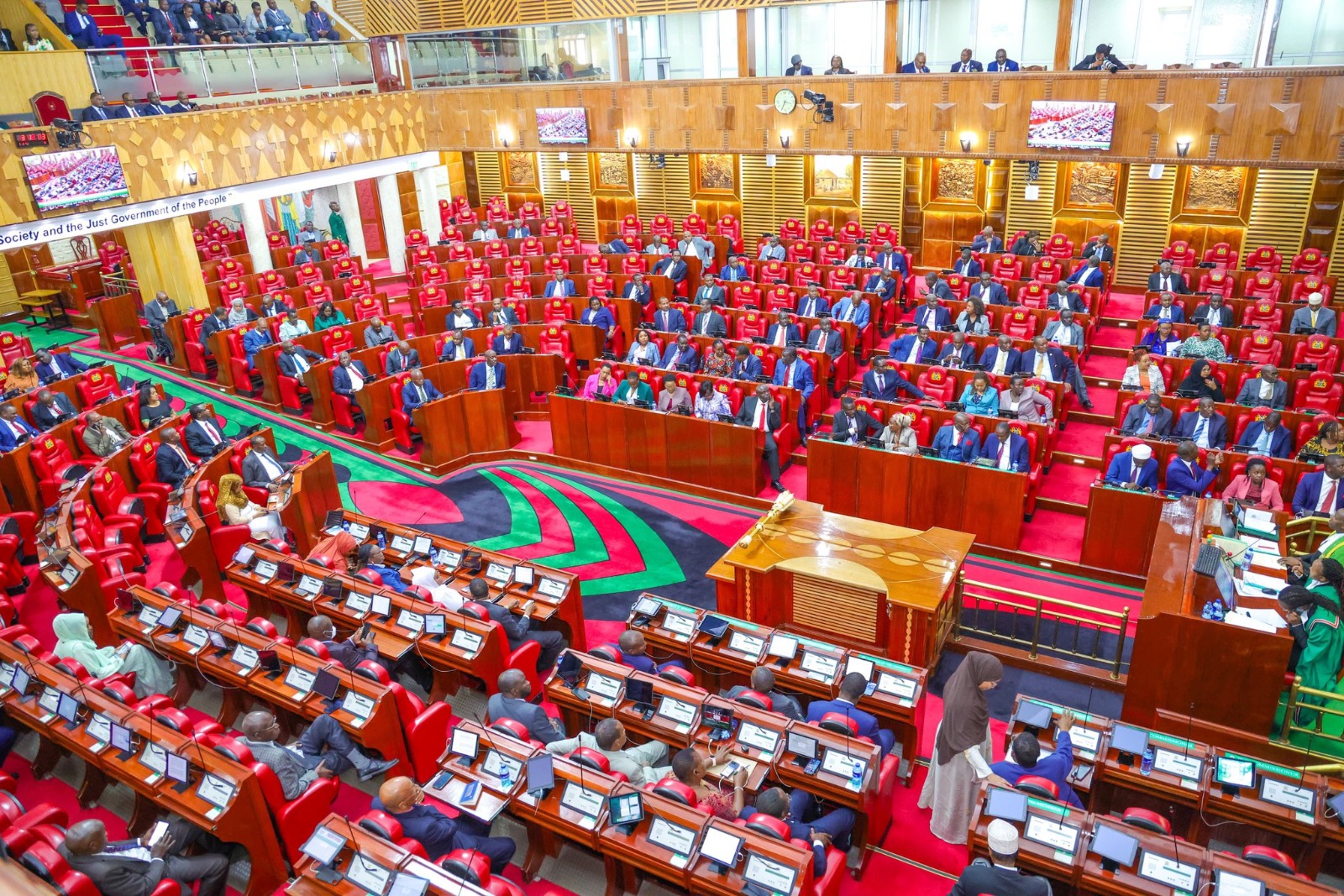 Parliament chamber filled with lawmakers seated in red chairs around wooden desks, with a central podium and colorful carpet.