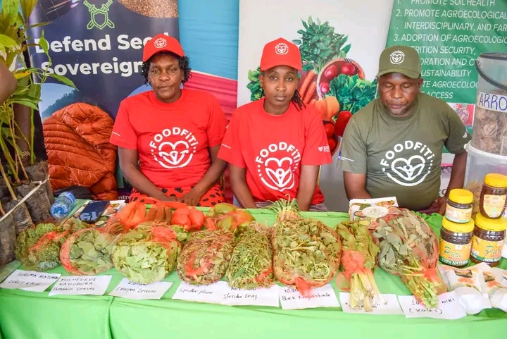 Three people in red FOODFIT shirts sit behind a table of fresh produce at a market stall.