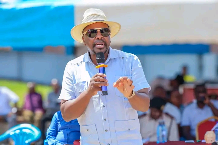 A man wearing a straw hat and sunglasses speaks into a microphone at an outdoor event, gesturing with his hand.