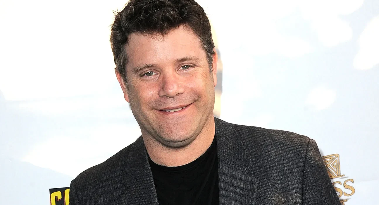 Smiling man in a dark blazer and black shirt at a red carpet or event backdrop San-friendly pose image.