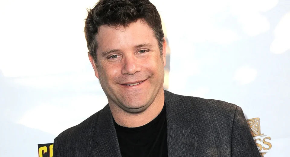Smiling man in a dark blazer and black shirt at a red carpet or event backdrop San-friendly pose image.