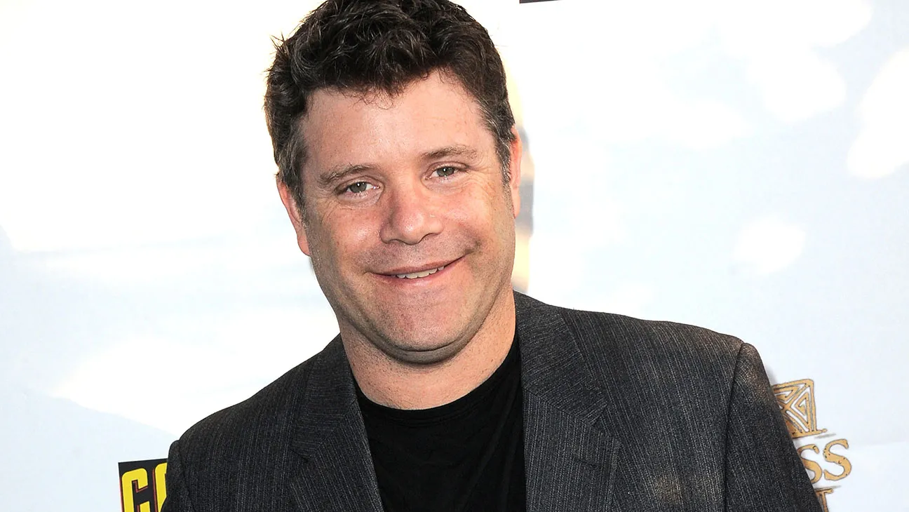 Smiling man in a dark blazer and black shirt at a red carpet or event backdrop San-friendly pose image.