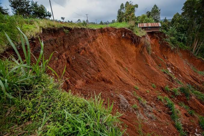 Red clay hillside eroded by a landslide, with green vegetation at the top and a small shed perched on the crest far right.