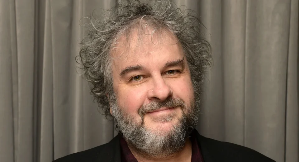 Smiling middle-aged man with gray hair and a salt-and-pepper beard, wearing a dark blazer in front of gray curtains.