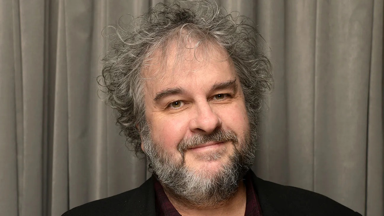 Smiling middle-aged man with gray hair and a salt-and-pepper beard, wearing a dark blazer in front of gray curtains.