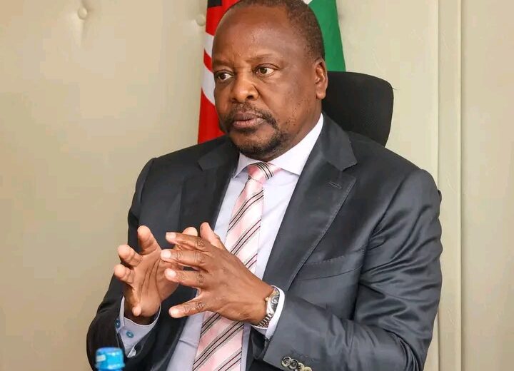 Professional man in a dark suit and striped tie seated in an office, gesturing with hands, a flag visible behind him.