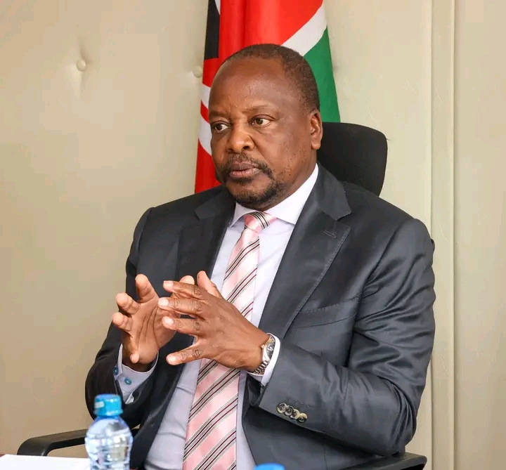Professional man in a dark suit and striped tie seated in an office, gesturing with hands, a flag visible behind him.