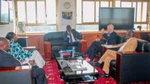 1786479611558 - Sacco Trend Magazine Six professionals in suits sit on leather sofas around a glass coffee table in a sunlit office, deep in discussion; water bottles and magazines on the table, a large TV screen on the wall in the background.