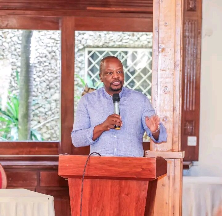 1787731567603 - Sacco Trend Magazine Man in a blue button-down shirt speaks into a handheld microphone at a wooden podium during a presentation in a room with wood paneling and windows.