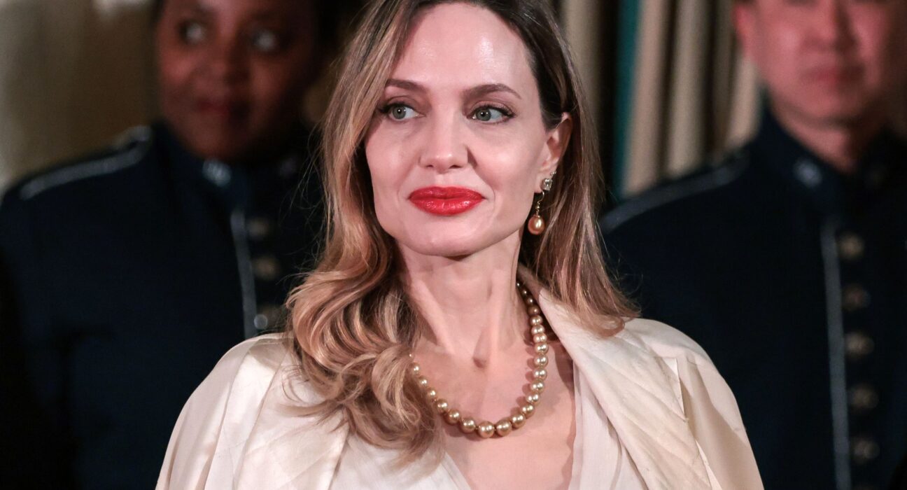 Angelina Jolie Net Worth - Sacco Trend Magazine Portrait of a woman with blonde hair, red lipstick, pearl necklace and a cream blouse, smiling slightly; blurred people in uniforms in the background.