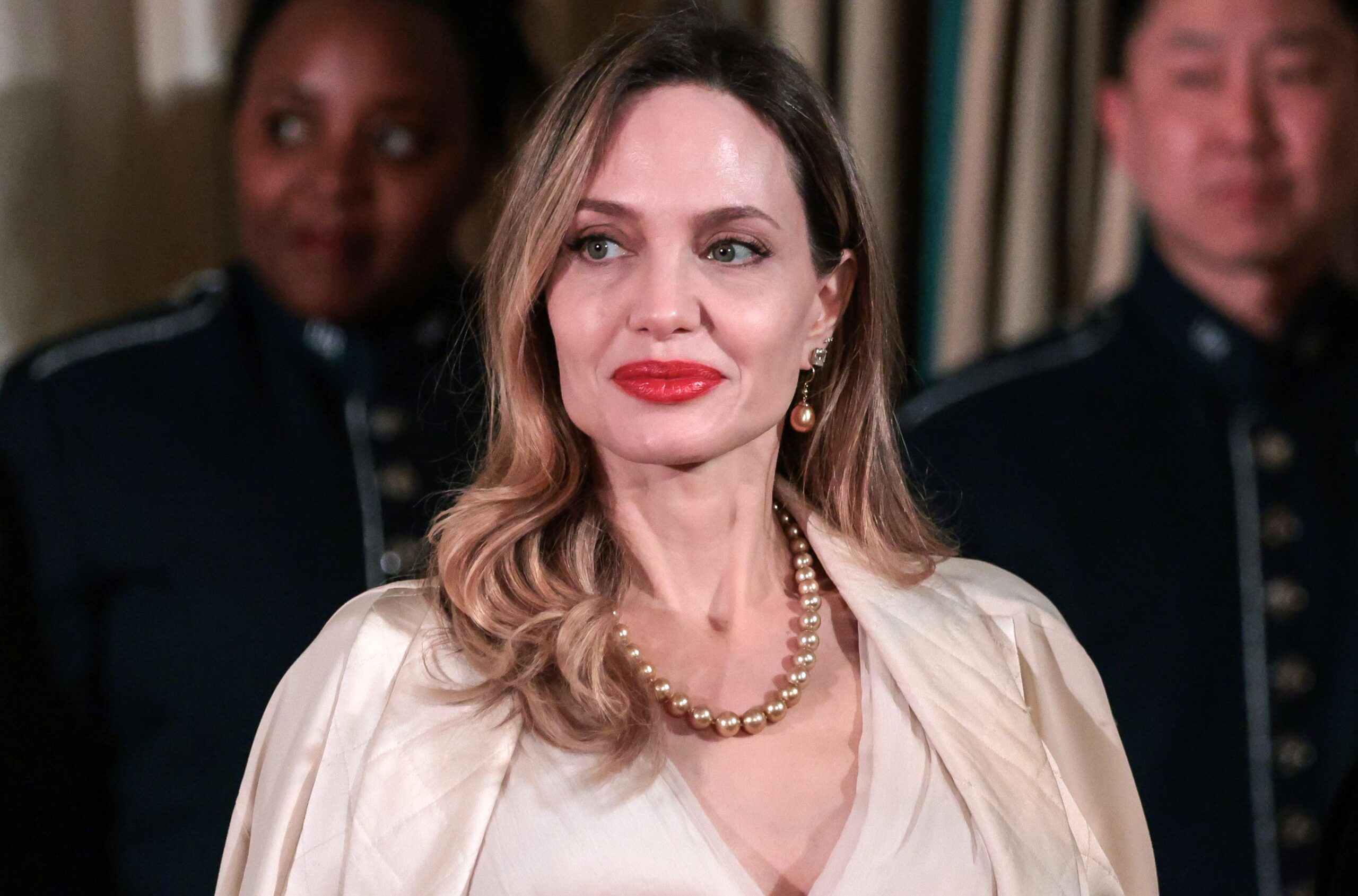 Portrait of a woman with blonde hair, red lipstick, pearl necklace and a cream blouse, smiling slightly; blurred people in uniforms in the background.