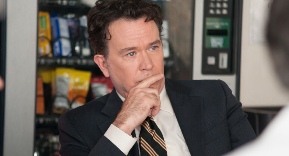 Man in a dark suit and striped tie rests his chin on his hand, appearing thoughtful in a blurred indoor setting