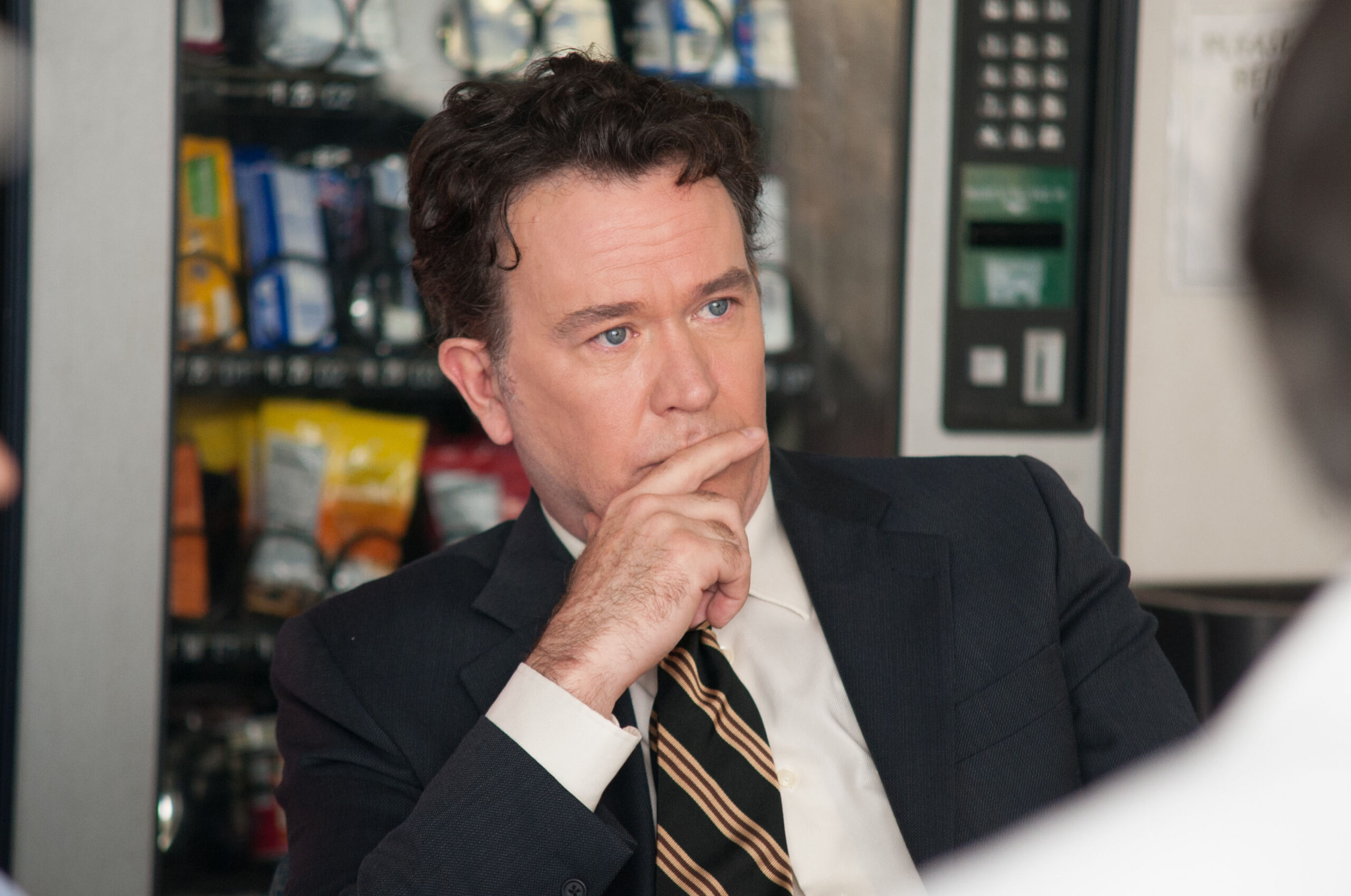 Man in a dark suit and striped tie rests his chin on his hand, appearing thoughtful in a blurred indoor setting
