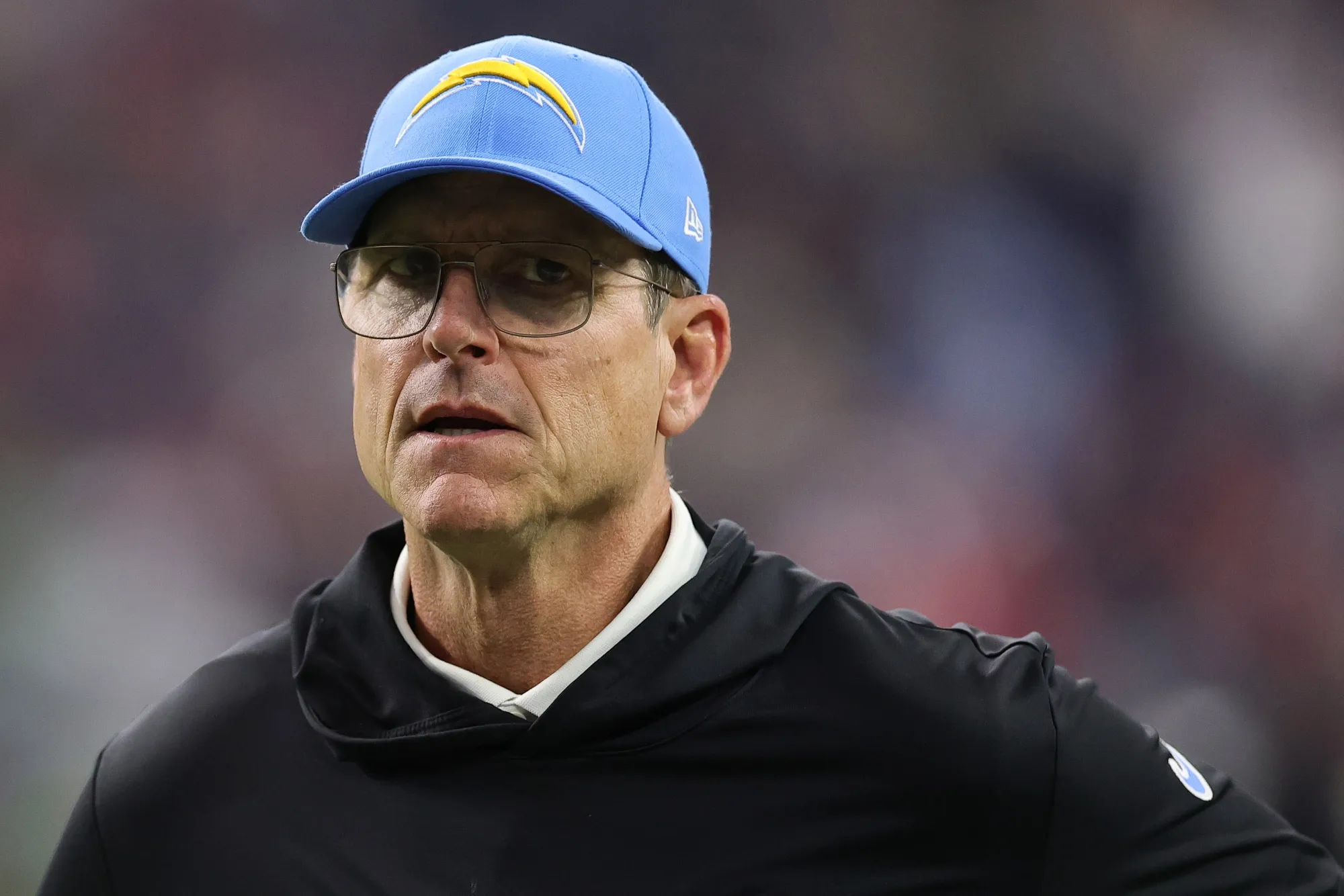 Close-up of a man wearing a light-blue Chargers cap and glasses, looking to the side in a stadium setting.