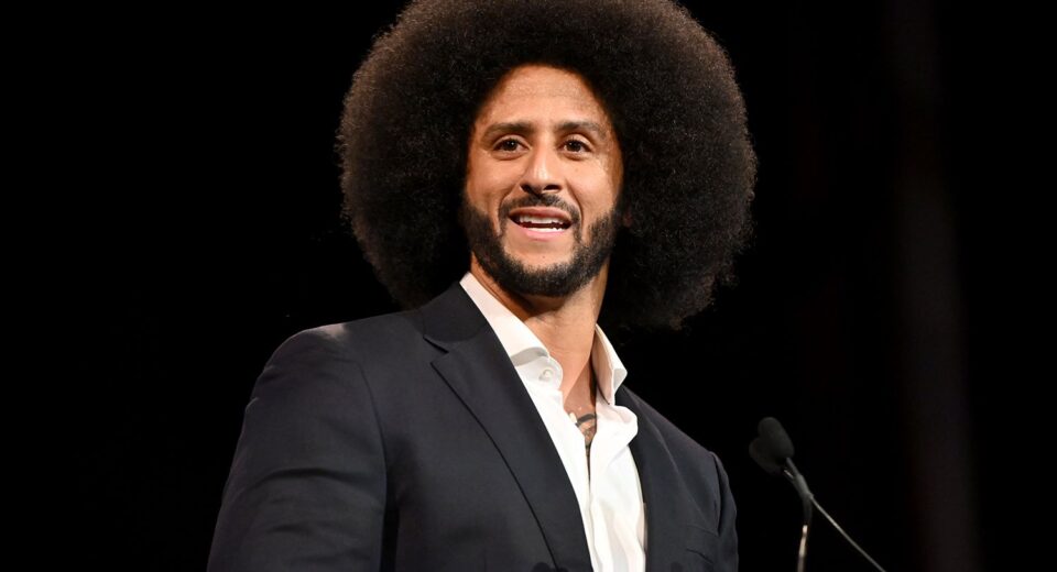 Man with a large Afro hairstyle delivering a speech at a podium in a dark background, wearing a navy suit and white shirt.