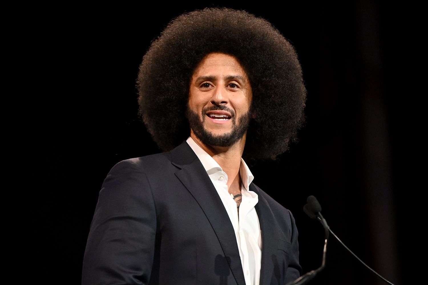 Man with a large Afro hairstyle delivering a speech at a podium in a dark background, wearing a navy suit and white shirt.