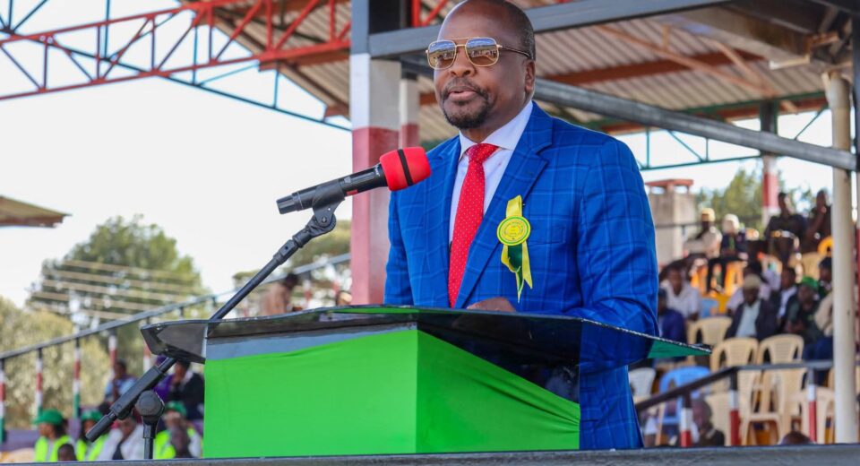Man in a blue checkered suit, red tie, and sunglasses speaks at a podium outdoors, wearing a yellow ribbon badge as an audience listens in the background.