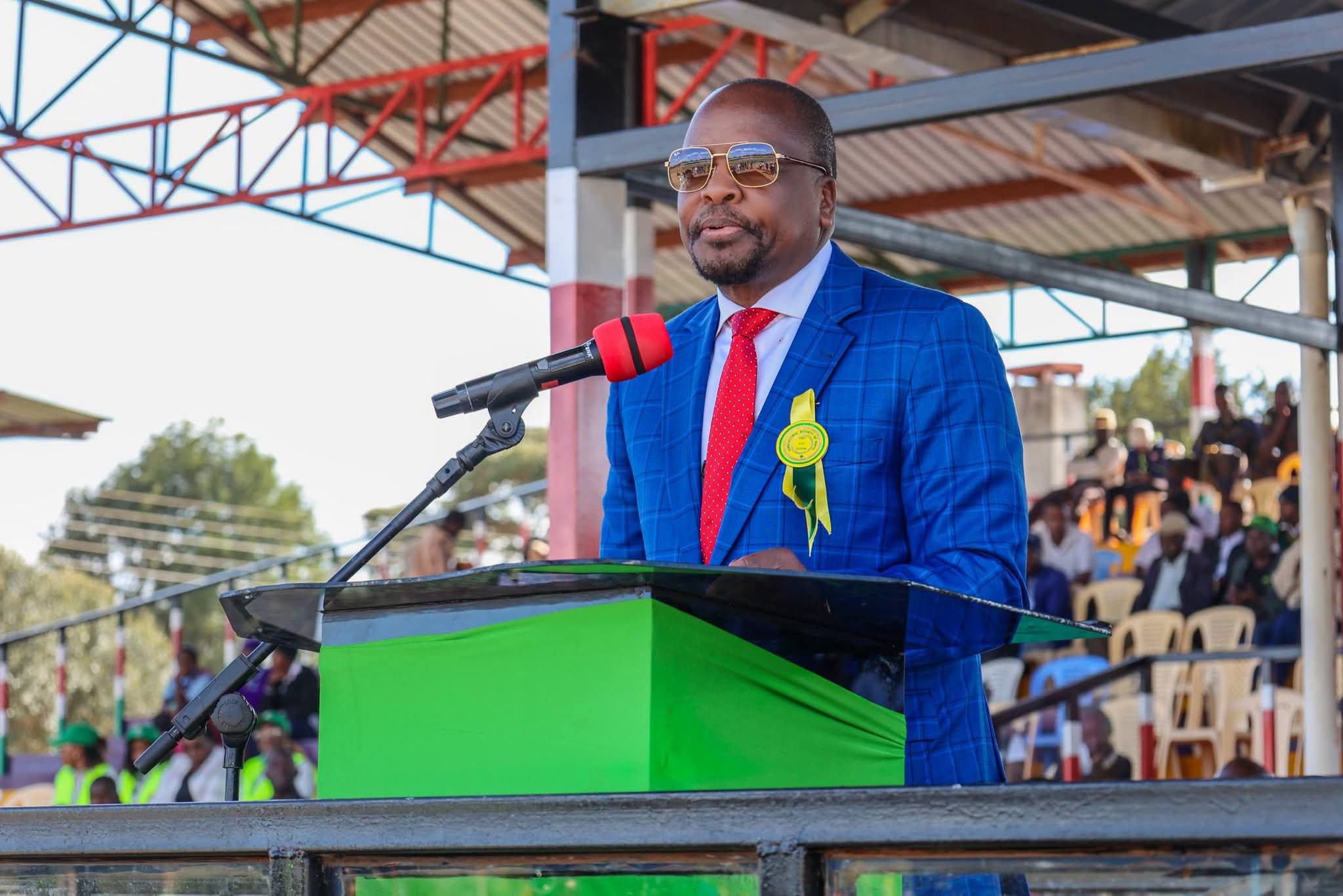 Man in a blue checkered suit, red tie, and sunglasses speaks at a podium outdoors, wearing a yellow ribbon badge as an audience listens in the background.
