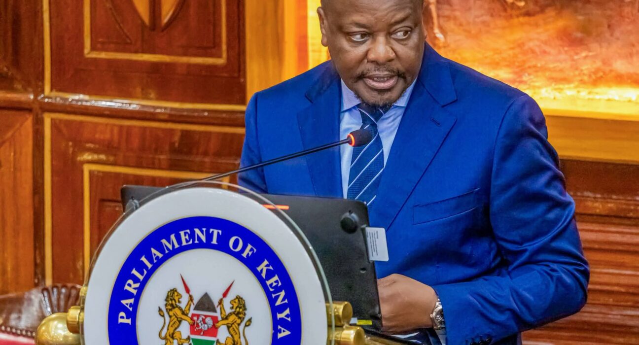 Man in a blue suit speaking at a podium with the Parliament of Kenya emblem on the front.