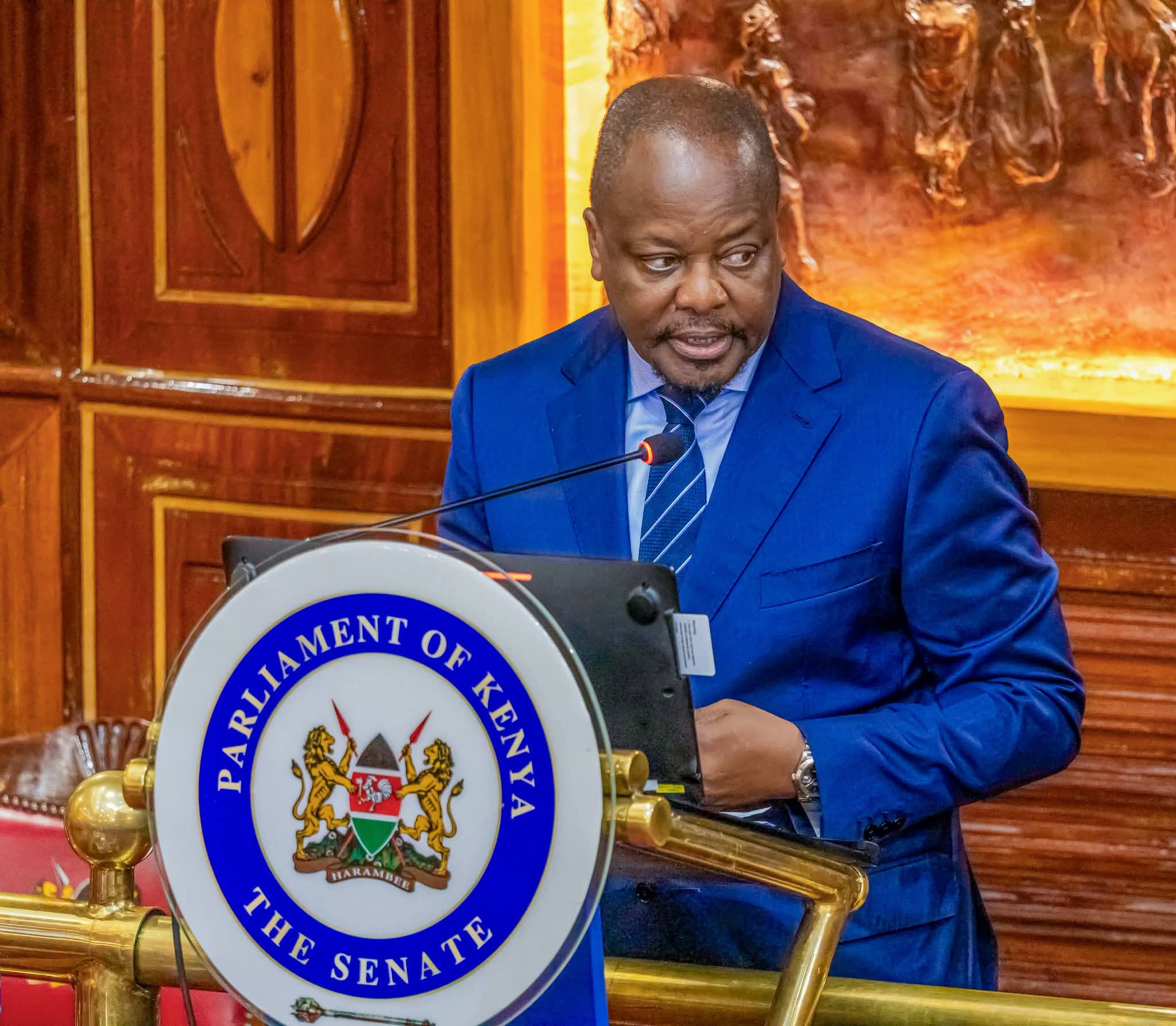 Man in a blue suit speaking at a podium with the Parliament of Kenya emblem on the front.