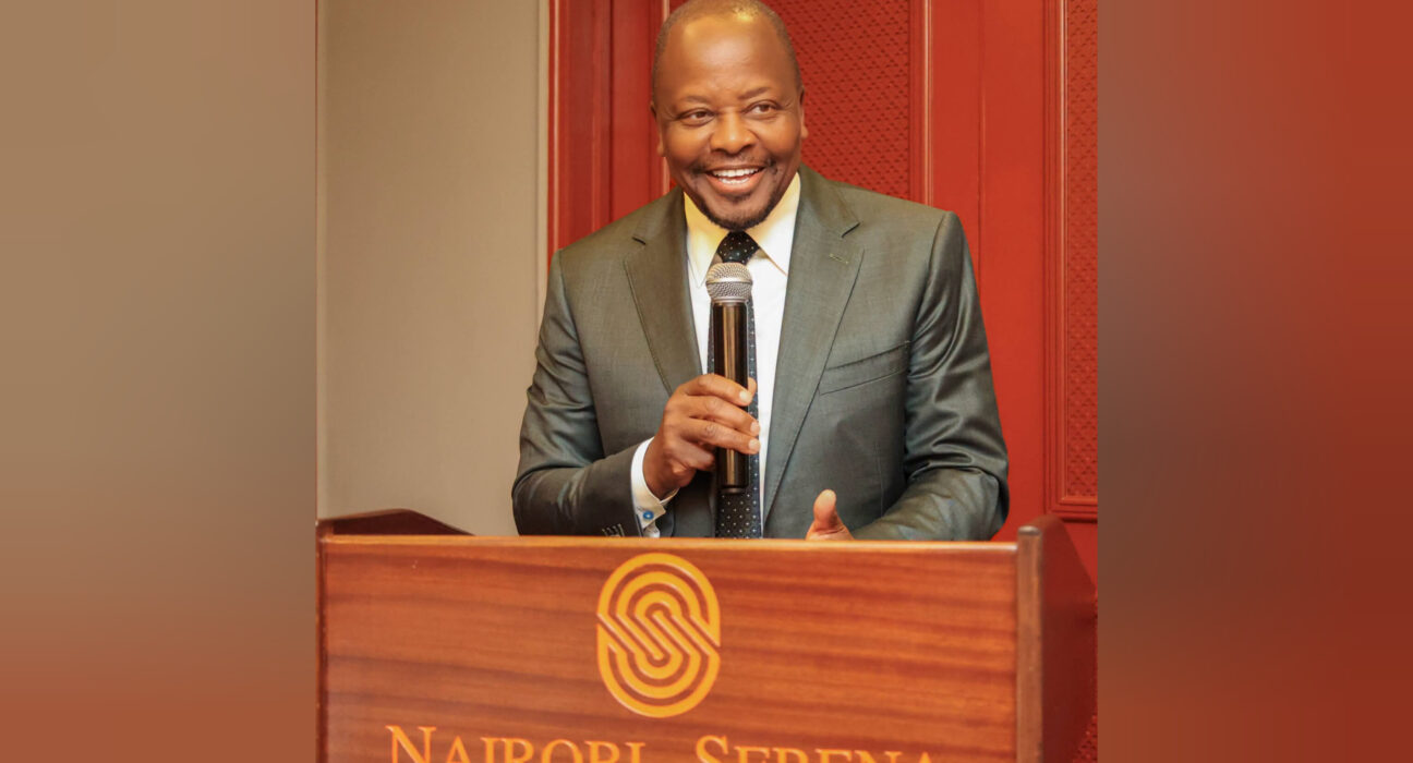 Man in a gray suit speaks into a microphone from a wooden podium with a Nairobi Serena logo on the front.