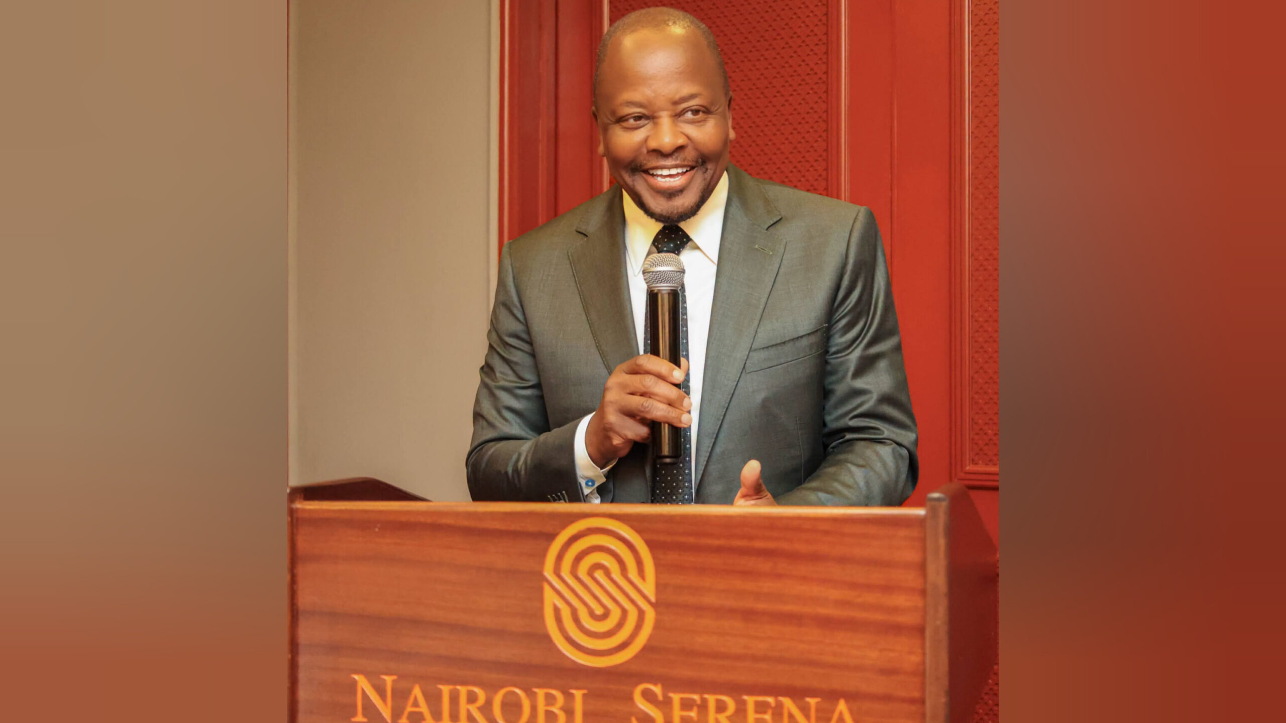 Man in a gray suit speaks into a microphone from a wooden podium with a Nairobi Serena logo on the front.