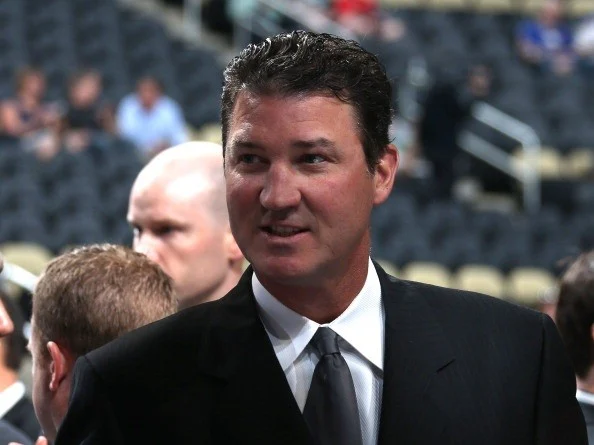 Man in a dark suit and tie at an event, with stadium seating in the background.