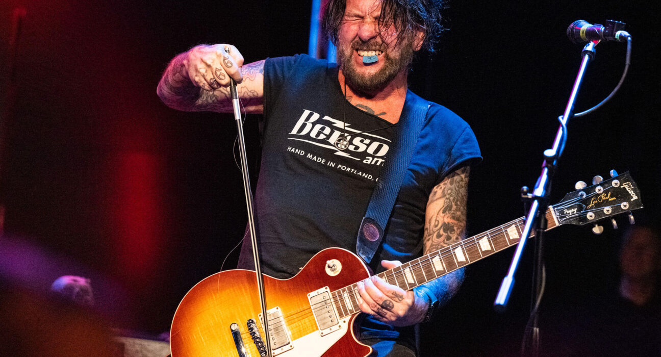 Male guitarist with a sunburst Gibson Les Paul performing on stage, gripping the mic stand with a tattooed arm visible.