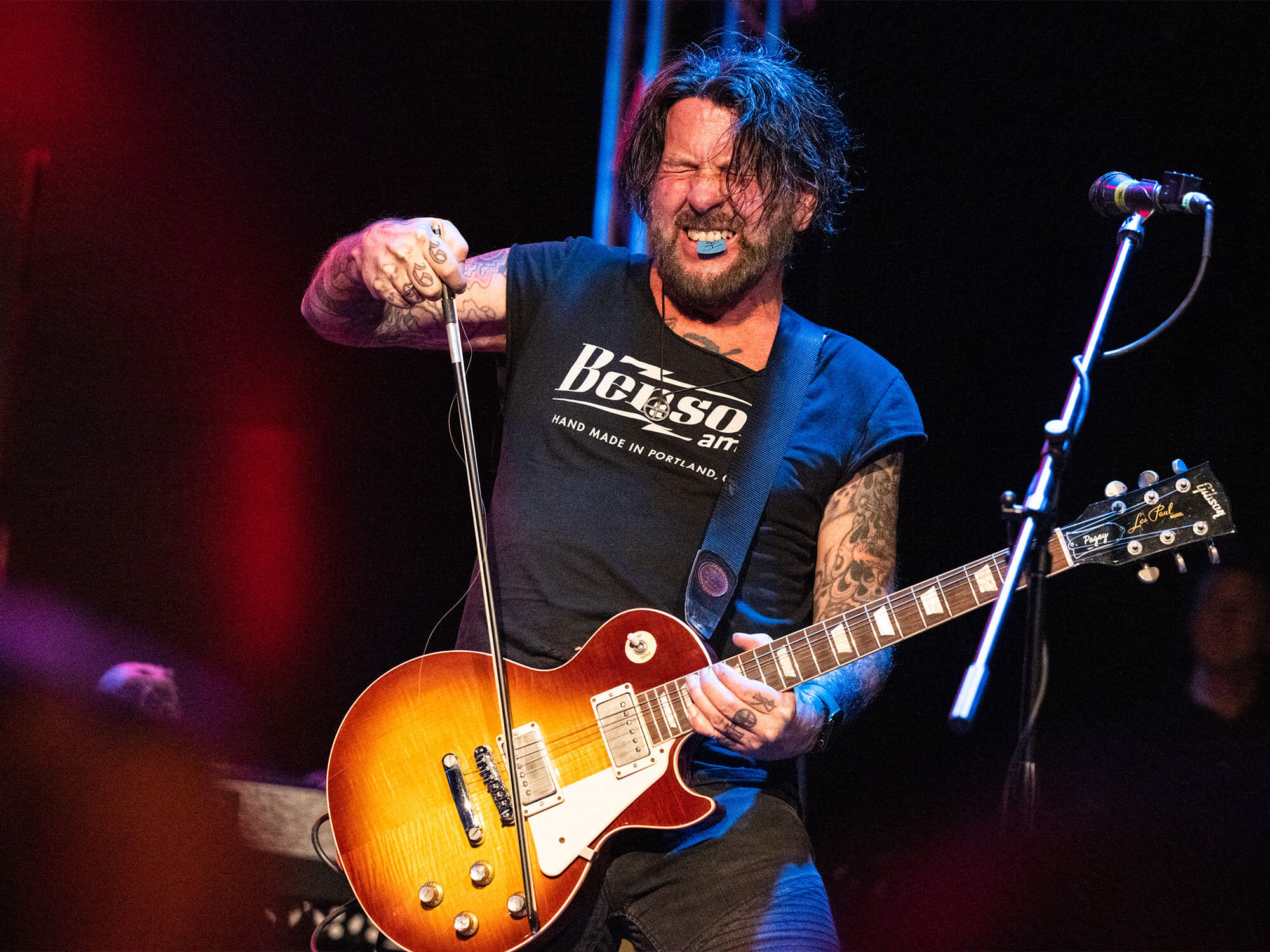 Male guitarist with a sunburst Gibson Les Paul performing on stage, gripping the mic stand with a tattooed arm visible.
