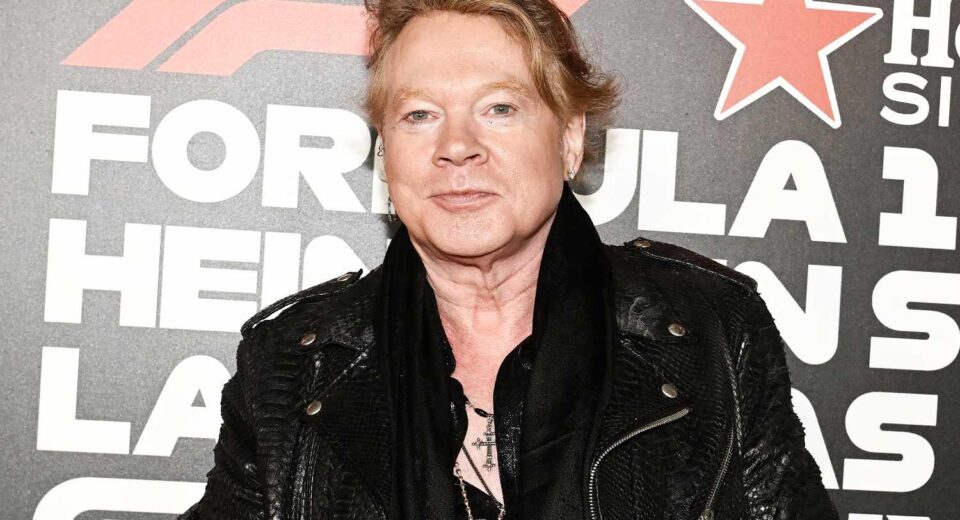 Middle-aged man with blond hair wearing a black leather jacket at a red-carpet event, standing in front of a backdrop with large white text and a red star motif.
