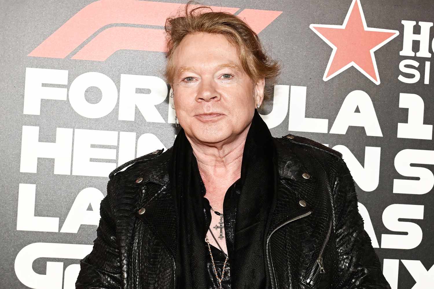 Middle-aged man with blond hair wearing a black leather jacket at a red-carpet event, standing in front of a backdrop with large white text and a red star motif.