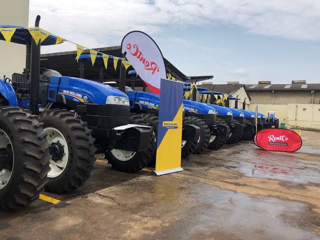 Row of blue New Holland tractors parked side by side at an outdoor display with yellow pennant flags overhead.