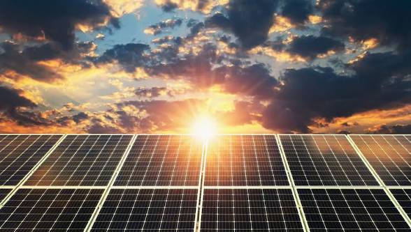 Sun setting behind a row of solar panels in a solar farm, with warm sky and sunburst between clouds.