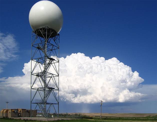 Tall radar tower with a white spherical dome against a blue sky and big cloud in an open landscape.