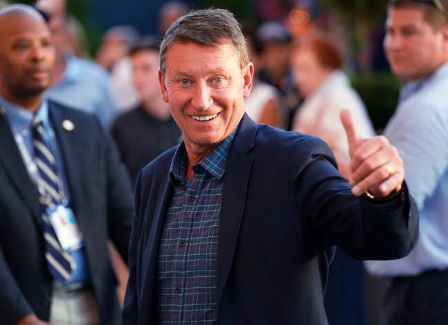 Smiling man in a navy blazer giving a thumbs up at an outdoor event with a blurred crowd behind him.