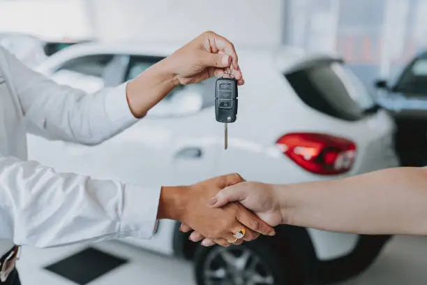 Person hands over car key while shaking hands in a car dealership showroom behind them a white car is visible.