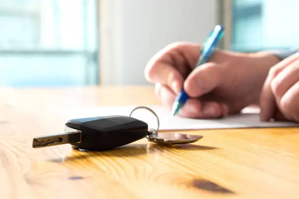 istockphoto-936987338-612x612 - Sacco Trend Magazine Close-up of a car key fob with a keyring on a wooden table as a person signs a document in the background.