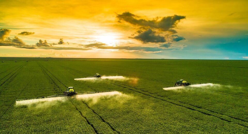 Wide field at sunset with three crop-dusting tractors spraying mist across rows?