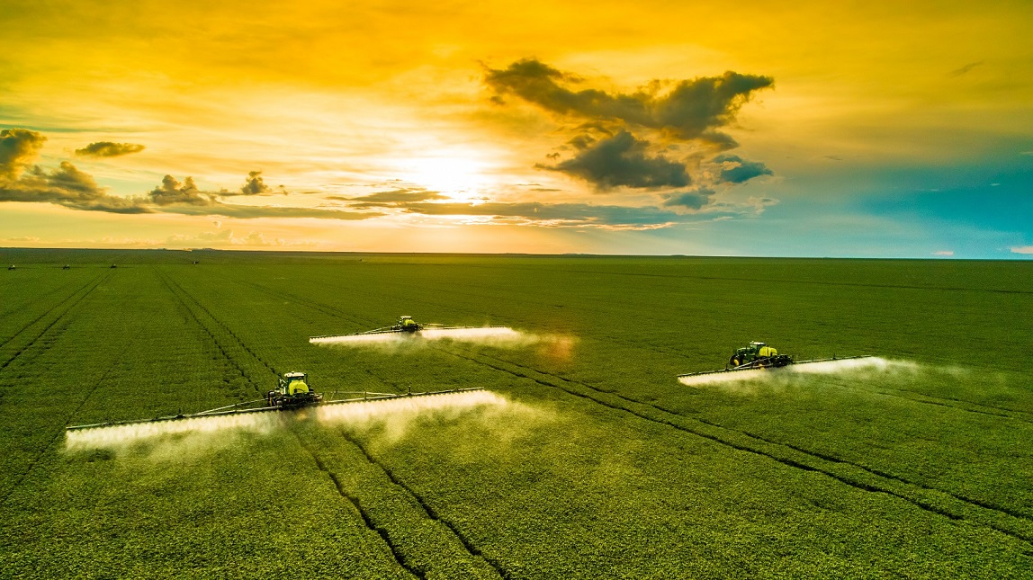 Wide field at sunset with three crop-dusting tractors spraying mist across rows?