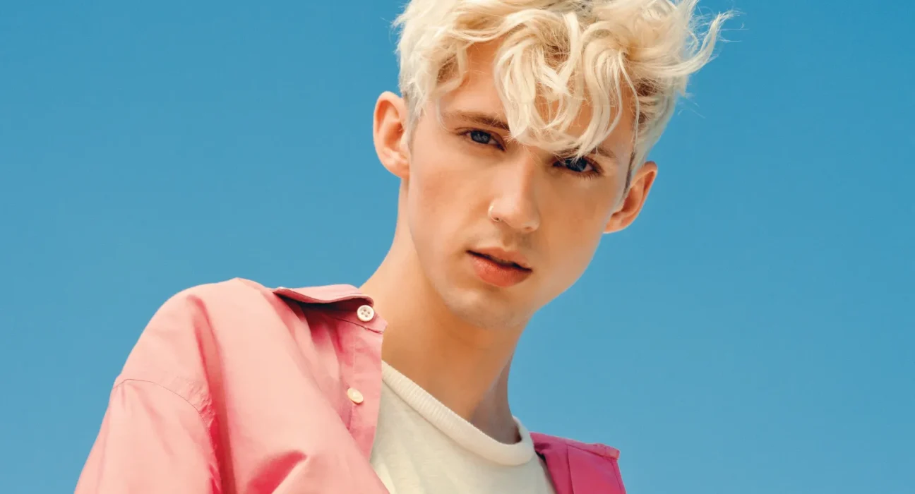 Portrait of a young man with platinum blond hair wearing a pink shirt over a white tee against a clear blue sky backdrop.