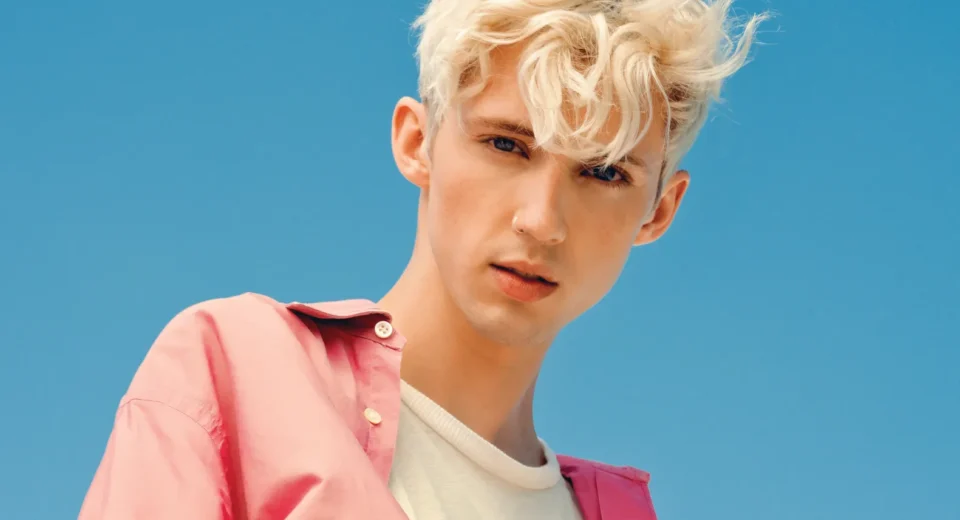 Portrait of a young man with platinum blond hair wearing a pink shirt over a white tee against a clear blue sky backdrop.